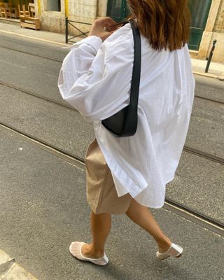 a stylish woman walking in a European city wearing an oversized white shirt, a small black shoulder bag, tan Bermuda shorts, and white mesh ballet flats