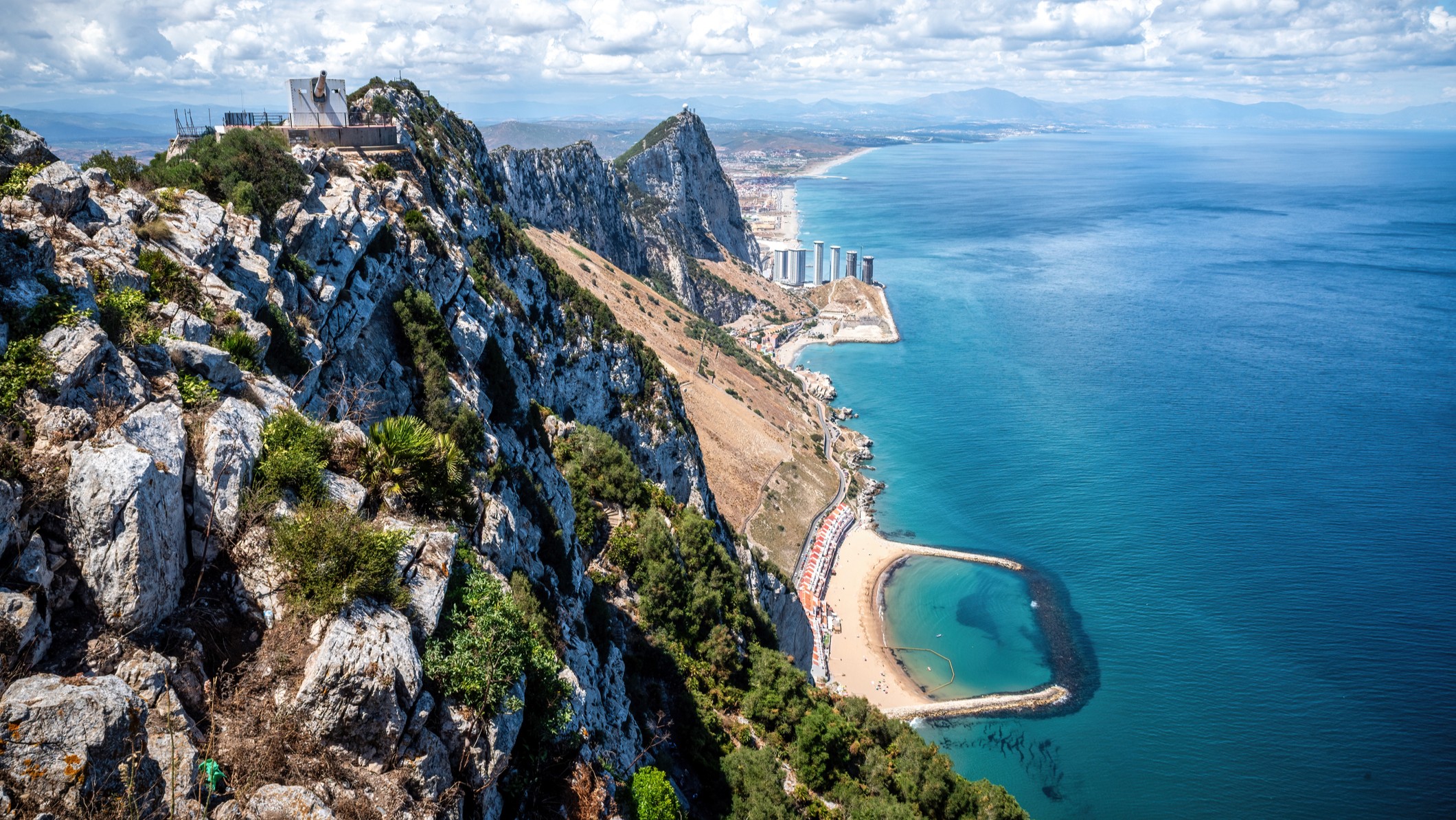 Aerial panorama capturing Gibraltar's dramatic limestone landmark, urban landscape, surrounding azure waters, and hazy African coastline under dramatic cloud formations