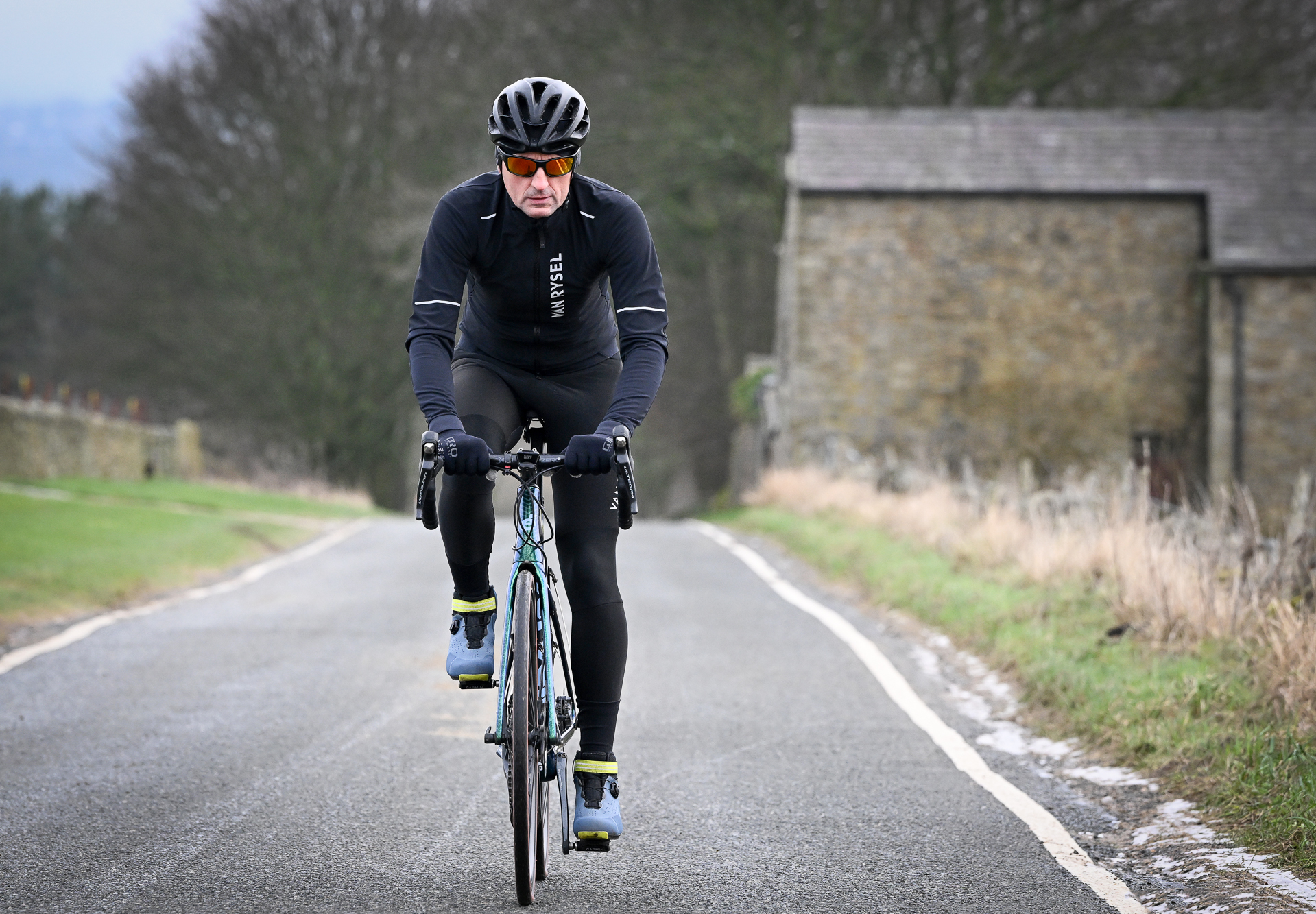 man riding towards the camera on a green road bike wearing a black jacket, black tights and blue shoes