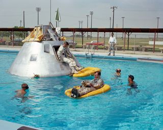 The prime crew of NASA's Apollo 1 mission practice water egress procedures in a swimming pool at Ellington Air Force Base in Houston, Texas in 1966. Astronaut Ed White rides a life raft in the foreground. Crewmate Roger Chaffee sits in hatch of the boilerplate model of the Apollo space capsule while Virgil "Gus" Grissom waits inside the spacecraft.