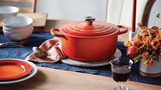 A Le Creuset cast iron dutch oven in orange on a table with an orange plate and orange flowers around it