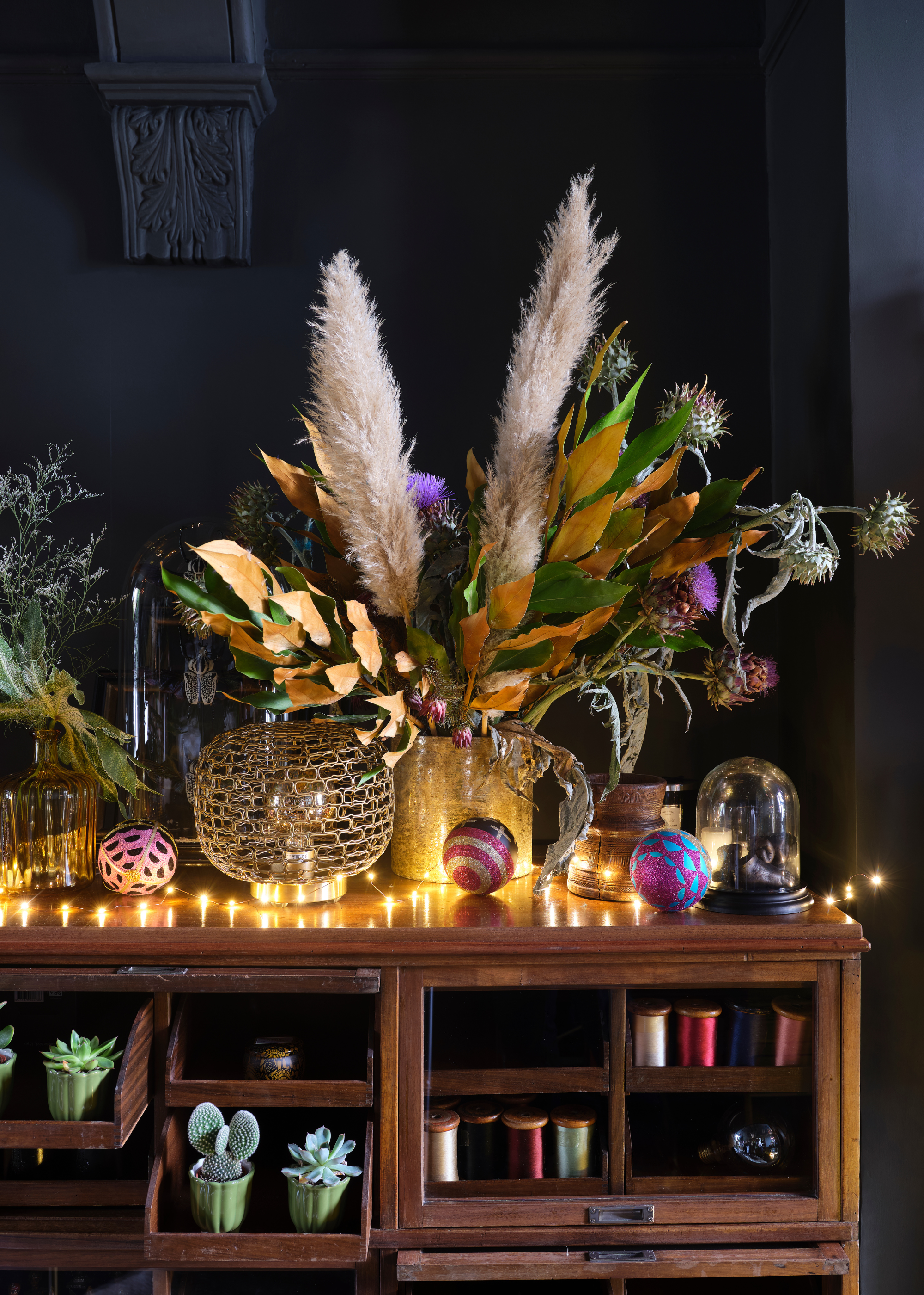 A cabinet is adorned with ornaments, baubles and foliage and is lit up with a set of string lights in front of a dark blue-painted wall