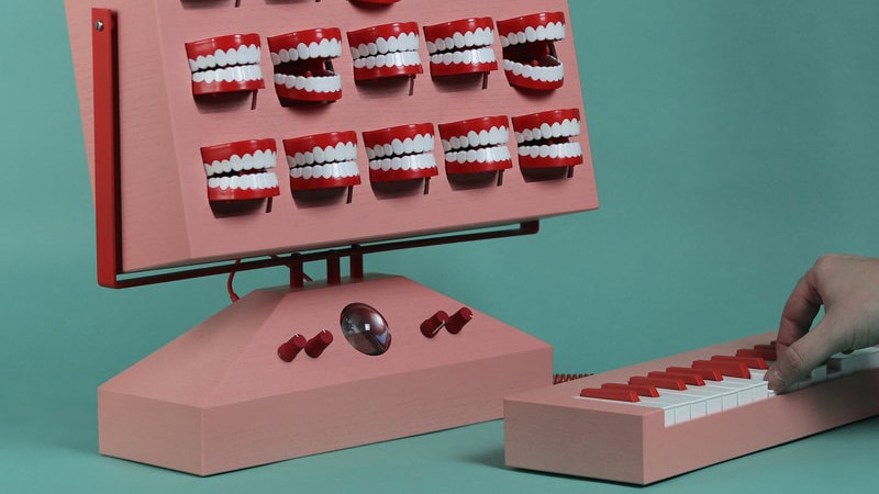 A hand plays a small, red-and-white keyboard that is connected to a larger, salmon-colored box with 25 chattering teeth toys mounted in a grid.