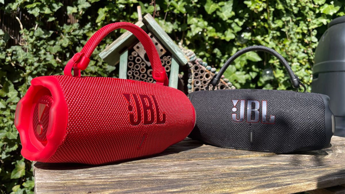 Two JBL Charge 6 wireless speakers (red on the left, black on the right) on a wooden table with a bush in the background.