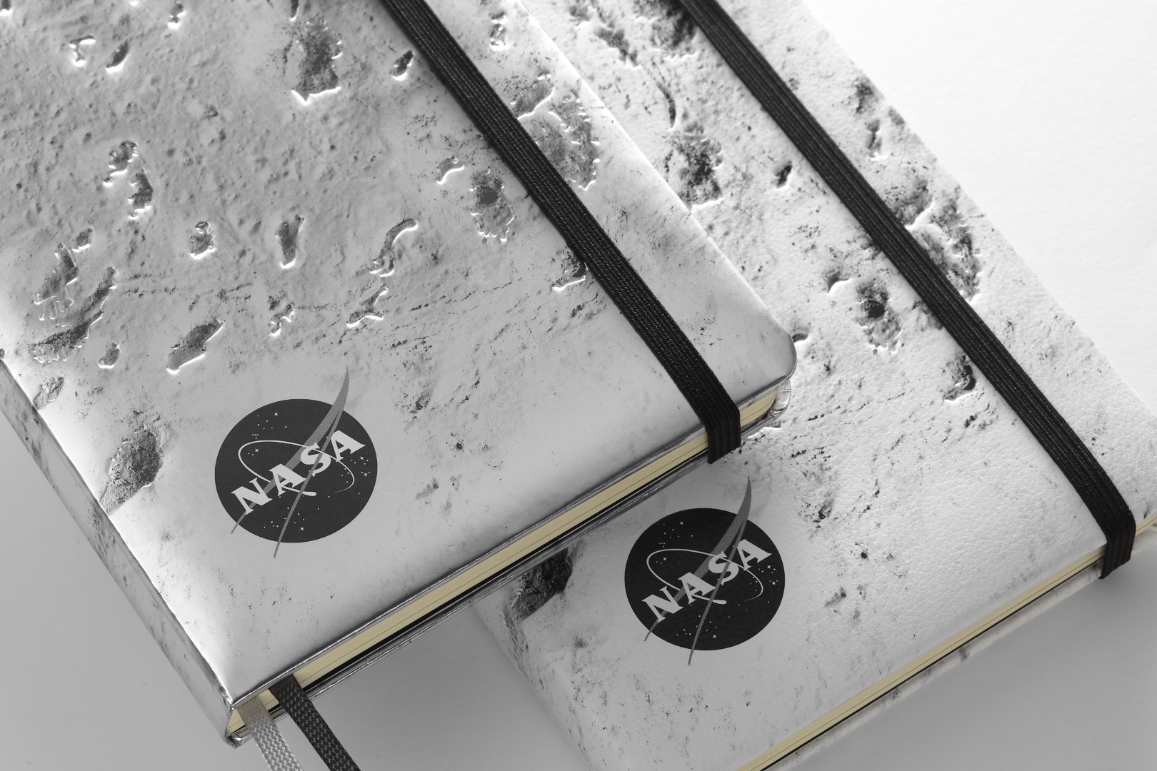 Two NASA-inspired Limited Edition Notebooks from Moleskine in dark silver and silver. Each cover is a close up of the moon's surface, has a black elastic closure, and a black and white version of the NASA logo is in the bottom left corner.