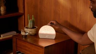 Hatch Restore 3 sunrise alarm clock on wooden bedside table next to cactus with man's arm reaching in to press button on top