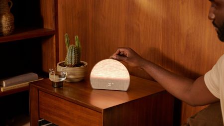 Hatch Restore 3 sunrise alarm clock on wooden bedside table next to cactus with man's arm reaching in to press button on top