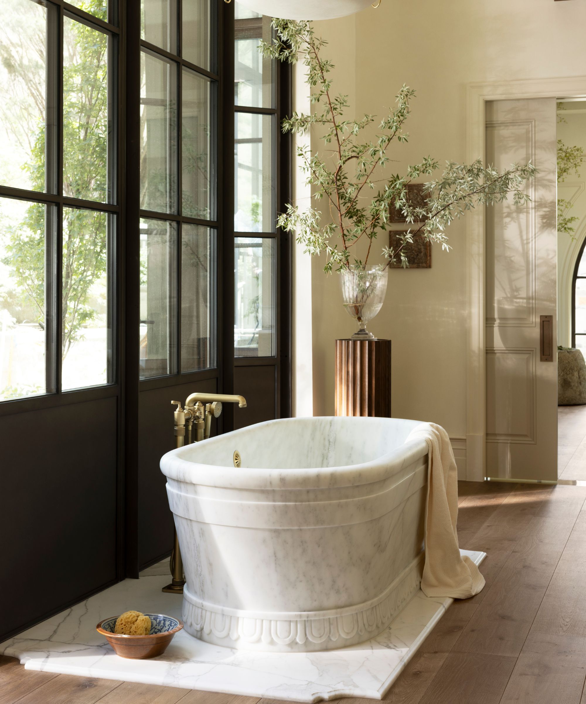 a neutral primary bathroom with large steel glass doors, a marble tub on a marble slab sits on wooden floors