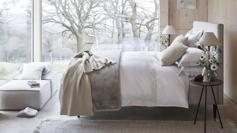 A pale grey and white bedroom with layered bedding on a double bed and a wintery scene visible through the bedroom's glass wall