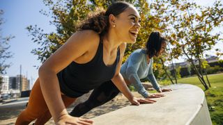 Two woman perform incline press-ups on a wall on a sunny day. Both are smiling and wearing fitness clothing