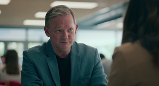 Daniel Lang (Douglas Henshall) sits at a desk, wearing a suit, looking at a woman sitting opposite him who has her back to the camera