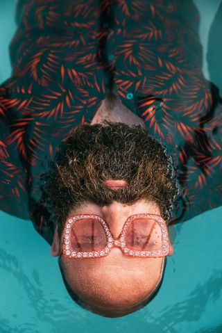 Person with sunglasses floating in water, wearing a tropical shirt