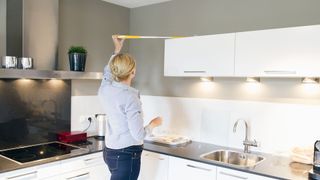 Woman cleaning the top of kitchen cabinets in a white kitchen to demonstrate a spring cleaning tip for apartments and small spaces