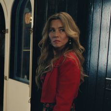 linda cardellini wears a red jacket and looks behind her while walking down a hall in no good deed