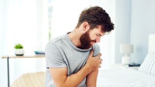 A man holding his shoulder in pain after waking up and sitting on the side of his bed