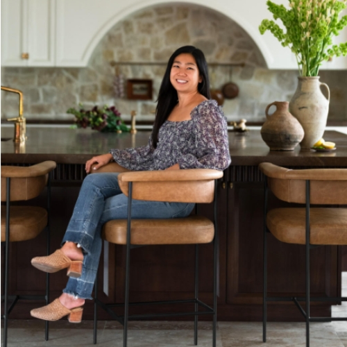 Picture of a woman in a flower blouse and jeans sitting in a kitchen at a bar stool.