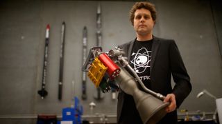 a man with curly hair, wearing a tshirt and sport coat, stands holding a small rocket engine. Small rockets hang on the unfocused grey wall behind him.