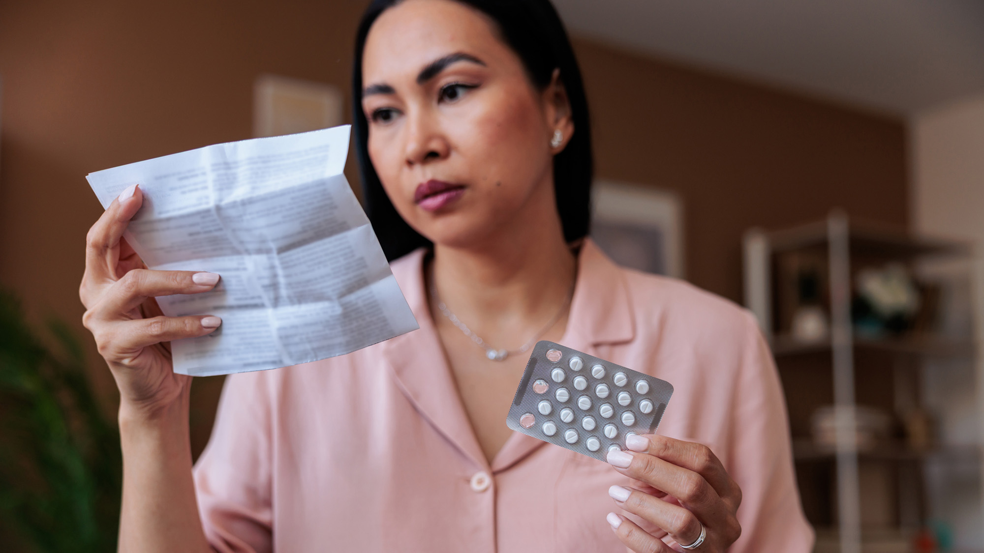 A woman with dark hair holds a blister pack of birth control and a piece of paper.