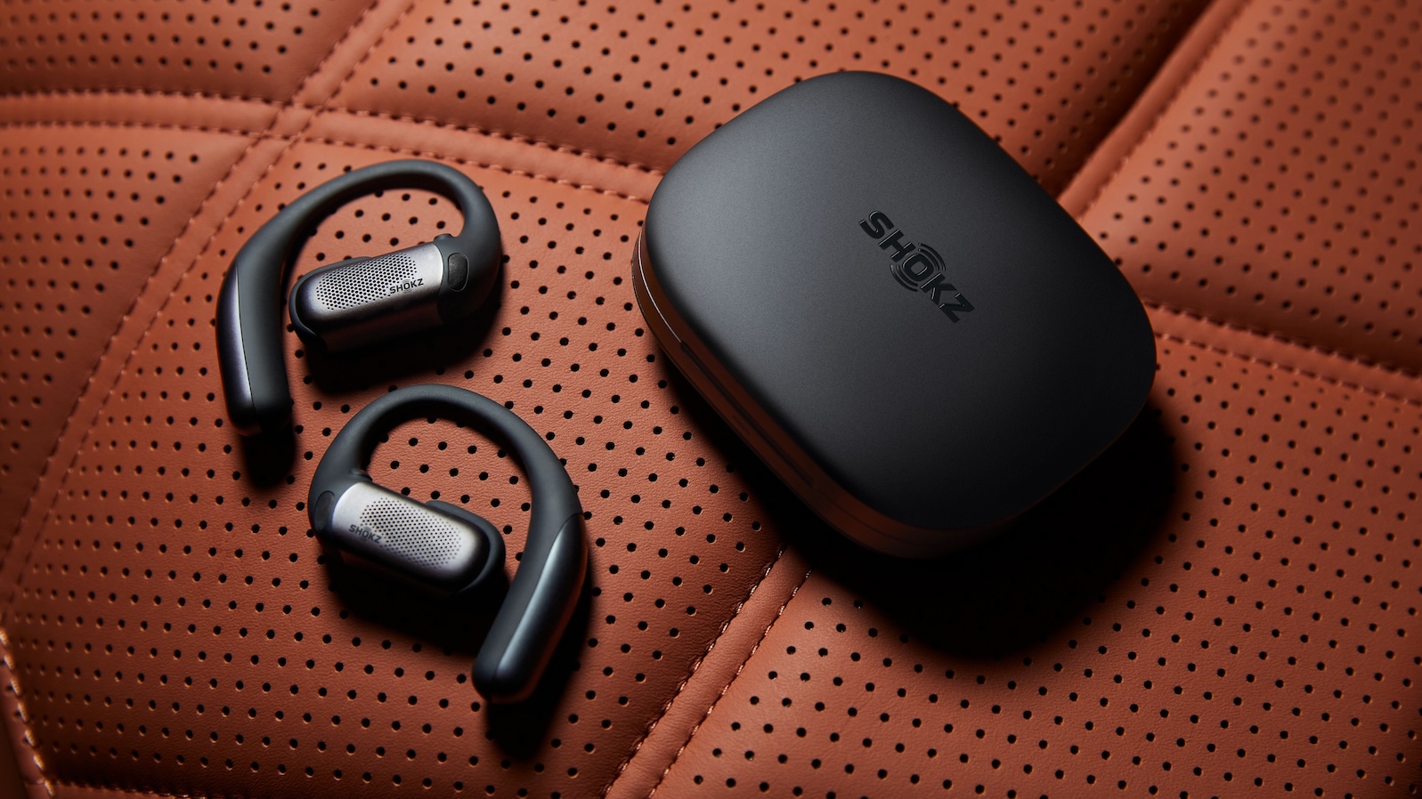 A black pair of Shokz OpenFit Pro earbuds with their carry case on a perforated orange surface.