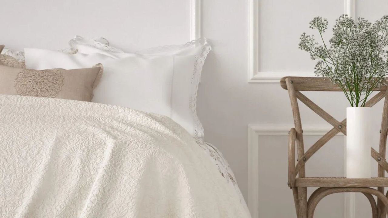 Cream Barefoot Dreams blanket on a bed against a white wall beside a wooden chair and a white vase.