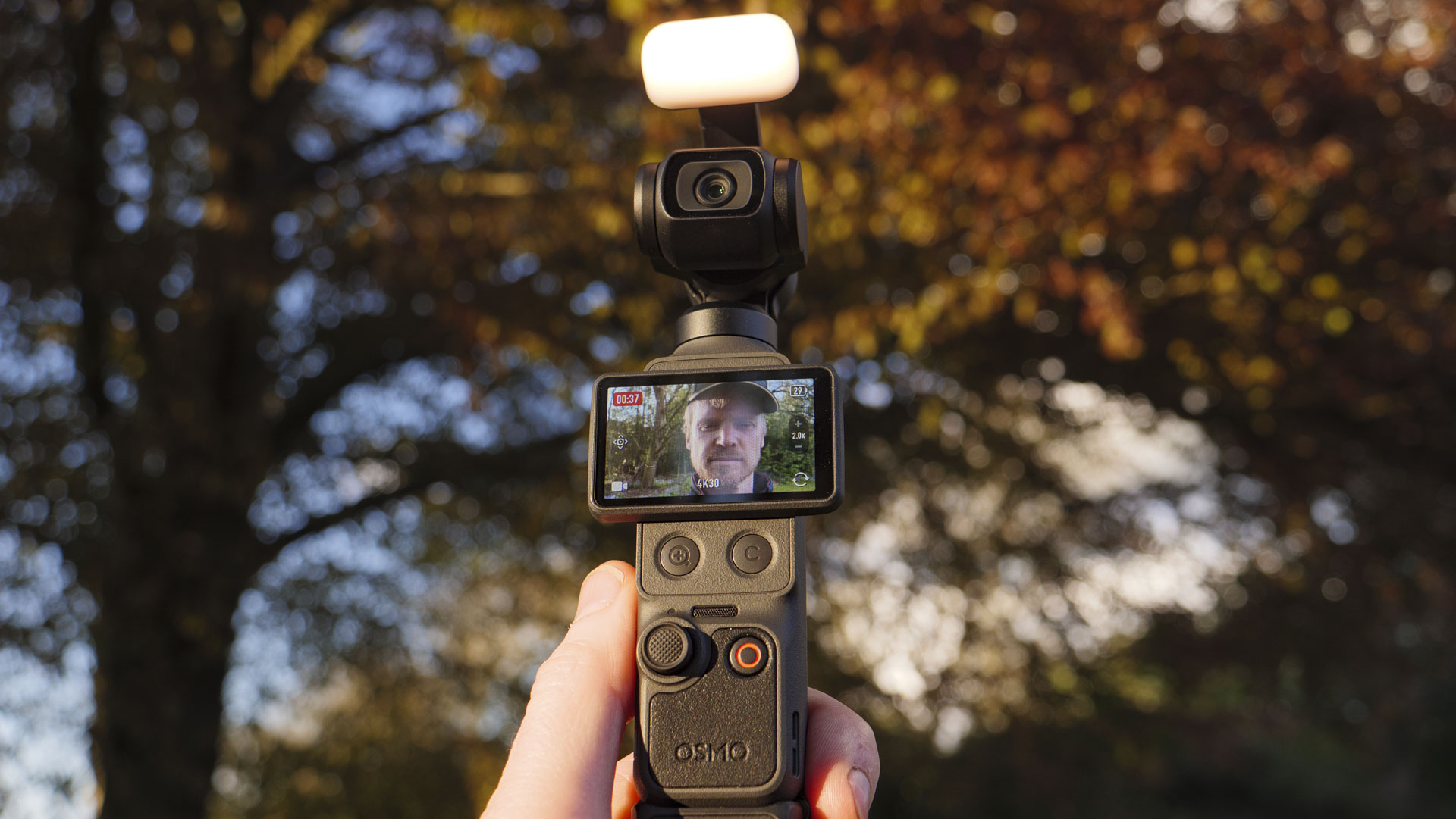 DJI Osmo Pocket 4 vlogging camera with natural background at golden hour, and the user in the frame of the screen