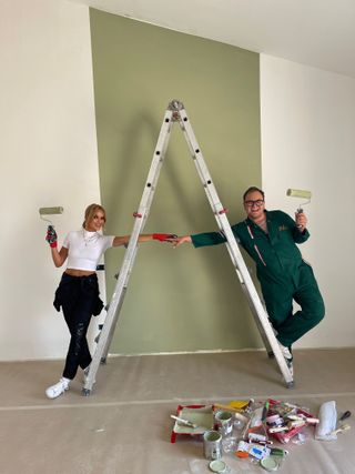 Amanda Holden and Alan Carr stand on either side of a stepladder, in front of an olive green feature wall. Each of them is holding a paint roller in one hand and reaching through the ladder with the other hand to hold hands