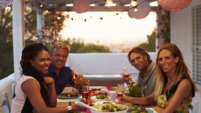 Two couples eat dinner on a porch at sunset. They are either in a shared vacation home or on a shared trip.