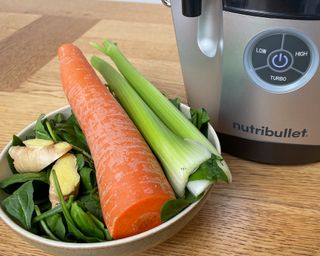 Nutribullet Pro Juicer on wooden dining table with whole unpeeled carrot, two stalks of celery, spinach leaves and two thumbs of ginger in ceramic bowl