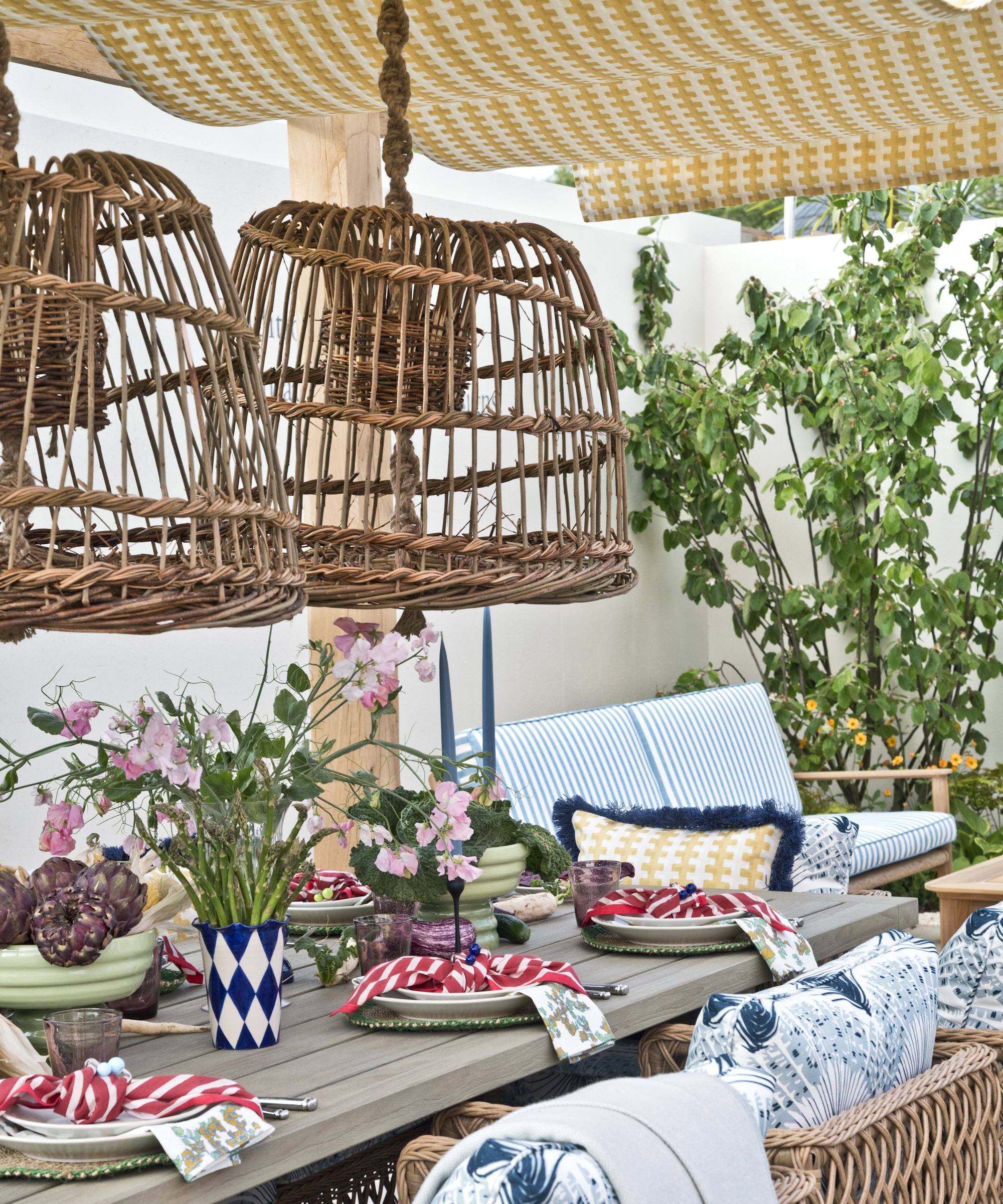 Patio dining table set with red napkins and sweet peas, with two large woven willow lamp shades above it