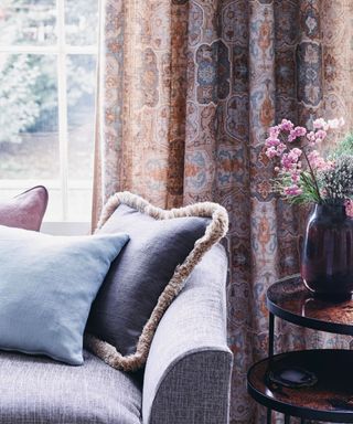 Living room with a window and floral patterned curtains, a sofa with cushions and side table.