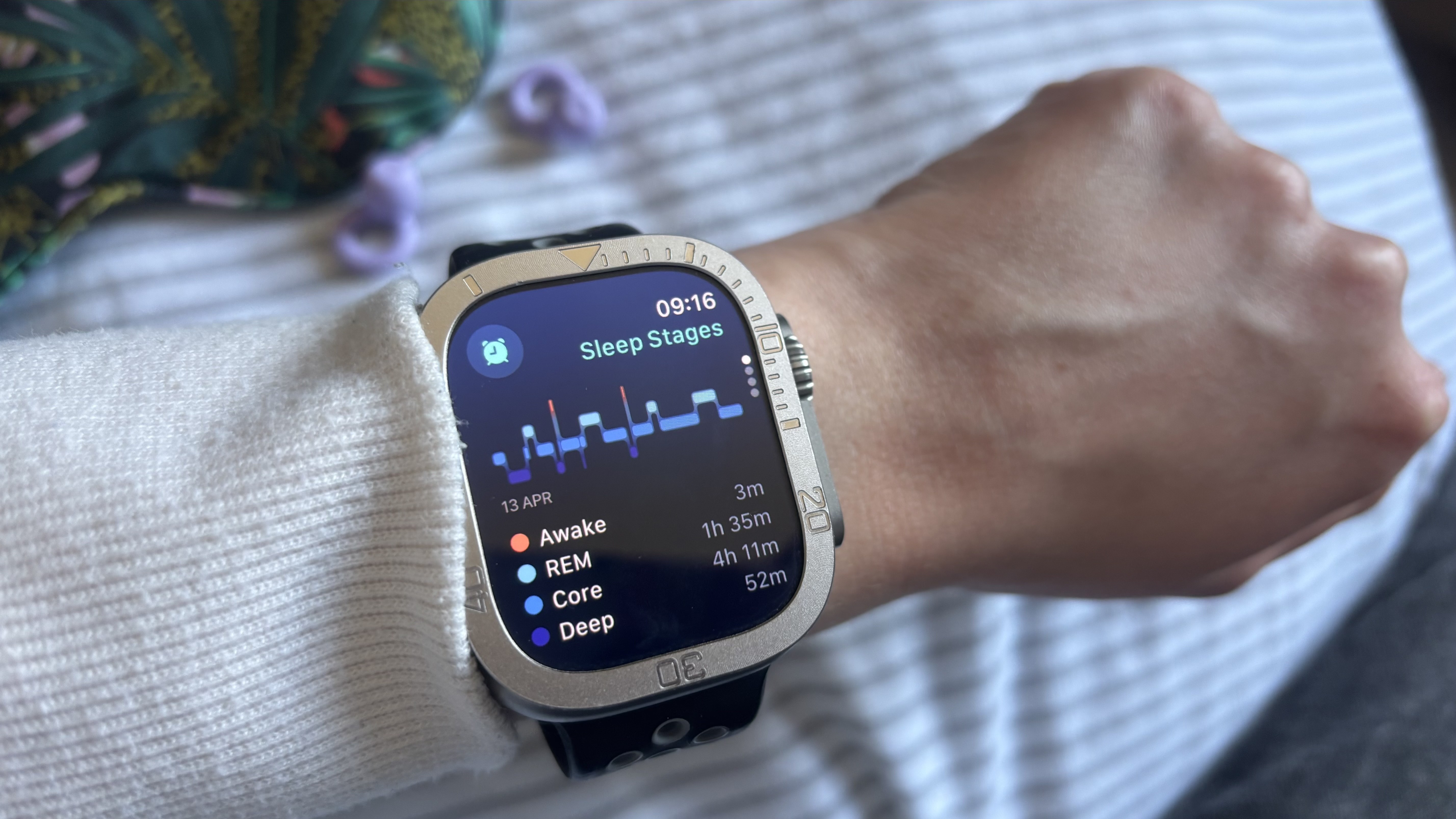 A close-up of the Apple Watch Ultra on a woman's wrist in a bedroom setting (a sleep mask and ear plugs are seen in the background). The screen displays Sleep Stages data for April 13th.
