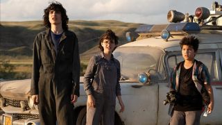 Finn Wolfhard, Mckenna Grace, and Logan Kim stand in front of Ecto-1 in a field in Ghostbusters Afterlife.