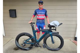 Dr Taylor stands behind his time trial bike dressed in red and blue kit wearing sunglasses and a cap