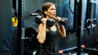 Woman holding two dumbells next to her shoulders, looking straight ahead at the gym