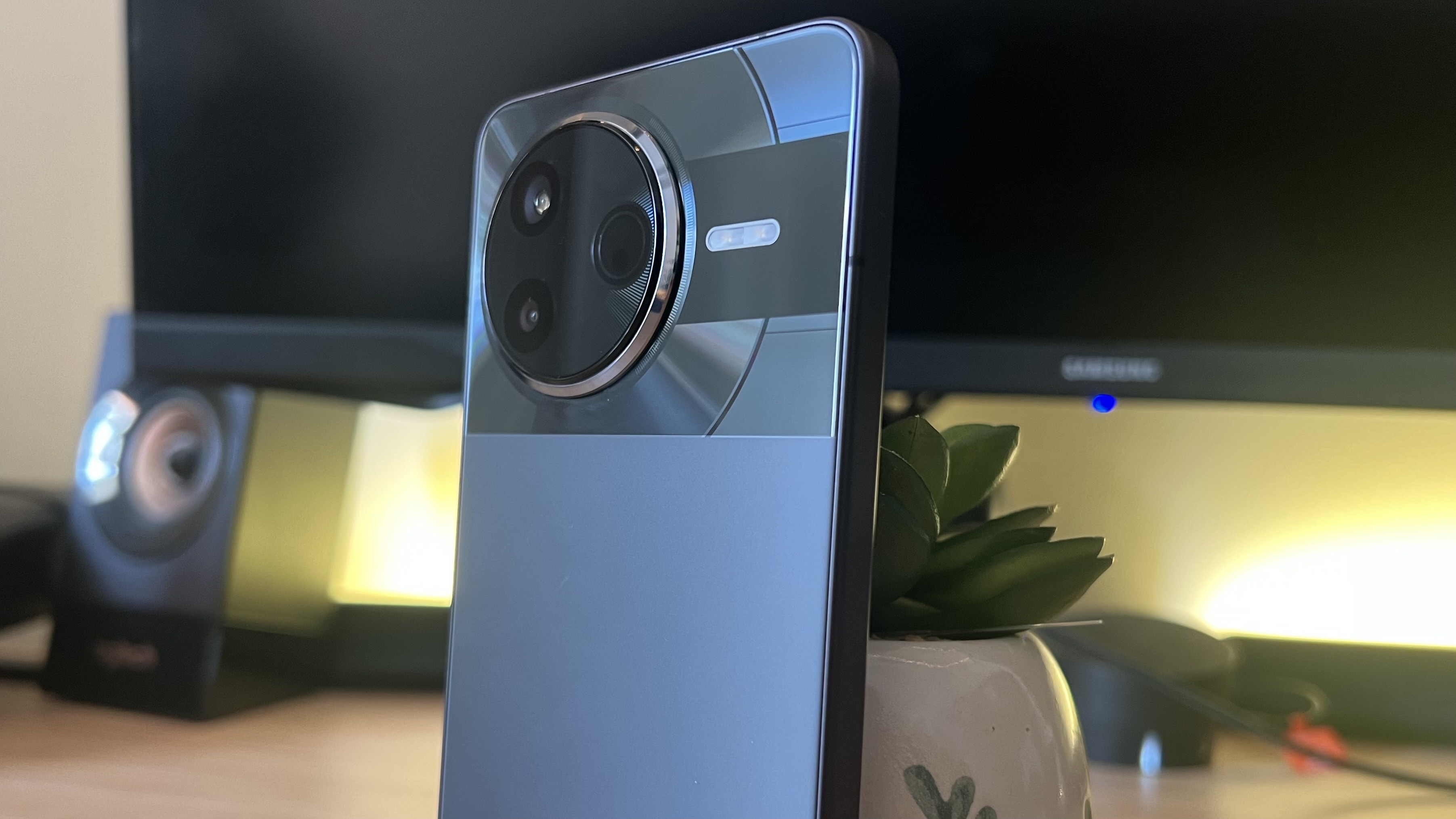 Poco F7 Pro smartphone camera bump and top third of rear panel leaning against a plant on a wooden desk