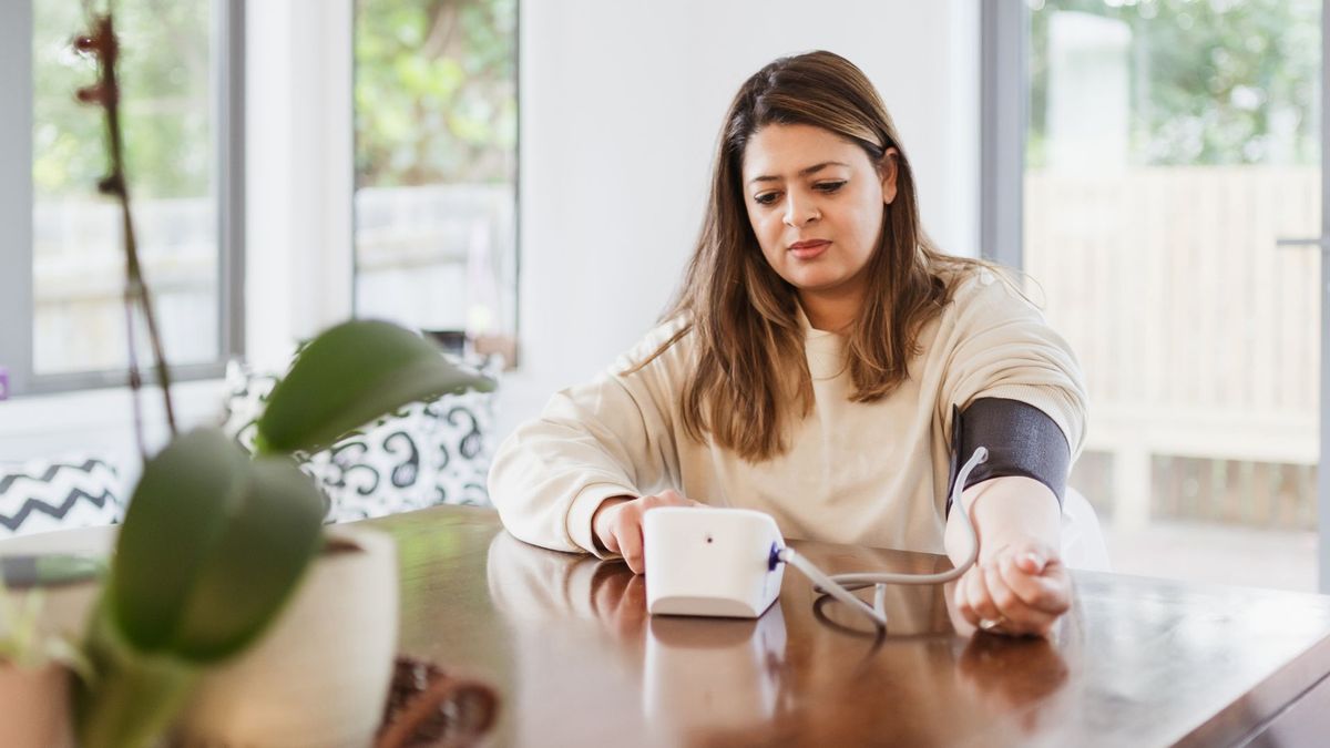 a woman with long brown hair sits at her kitchen table and uses an at-home blood pressure monitor, which includes an inflatable cuff on her arm