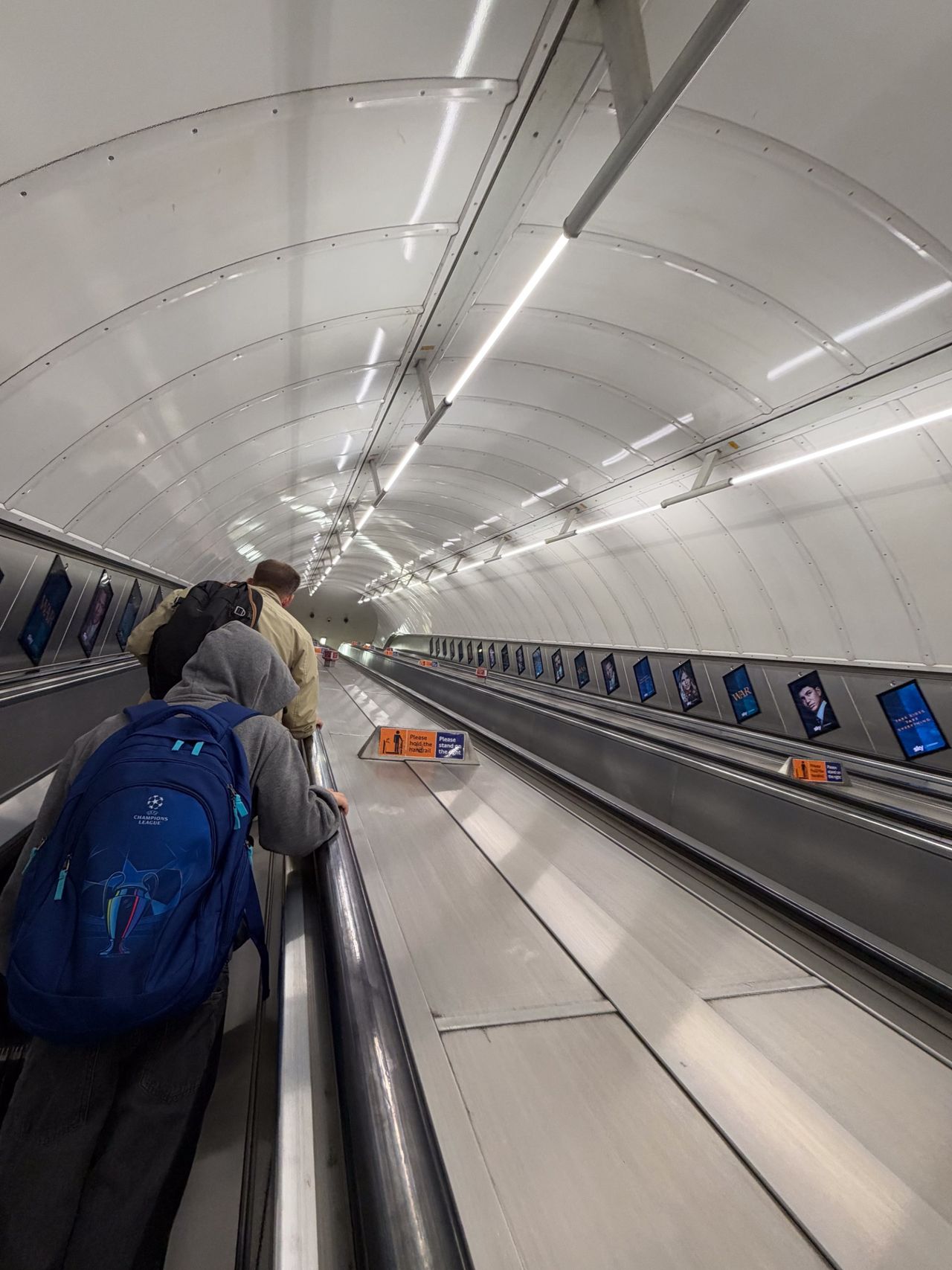 People riding a long moving walkway through a brightly lit curved tunnel on the London Underground.
