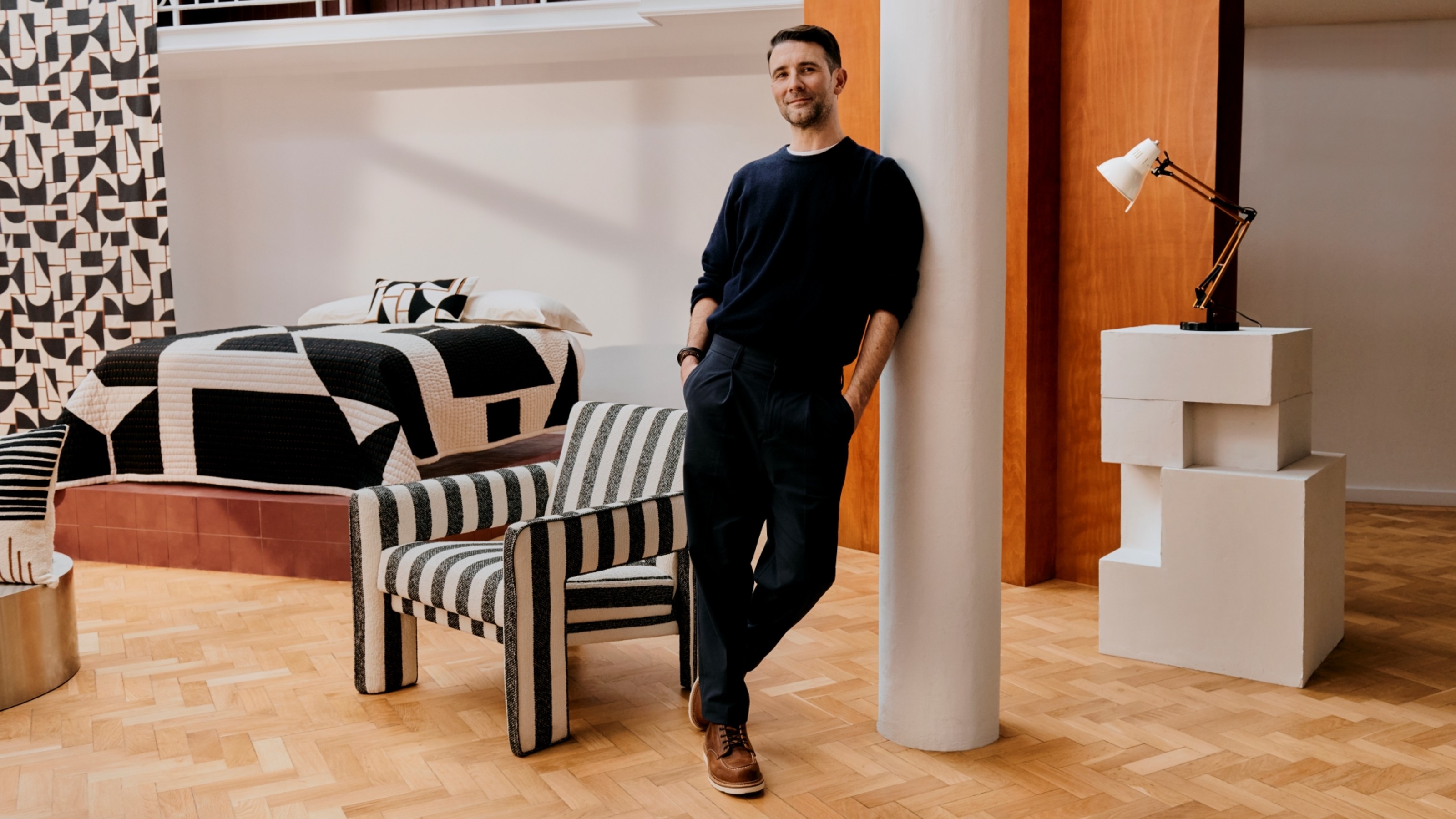 Interior Design Masters series 6 winner, John Cooper leaning against a pillar, surrounded by pieces from his John Lewis collection