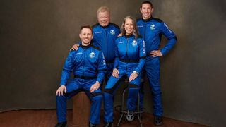 William Shatner and fellow NS-18 crew members in Blue Origin flight suits in front of a brown backdrop