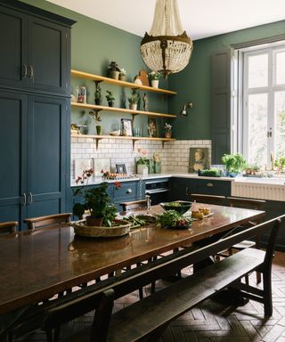 large wooden dining table in centre of blue kitchen with bench seating on one side and ornate chandelier hanging above table