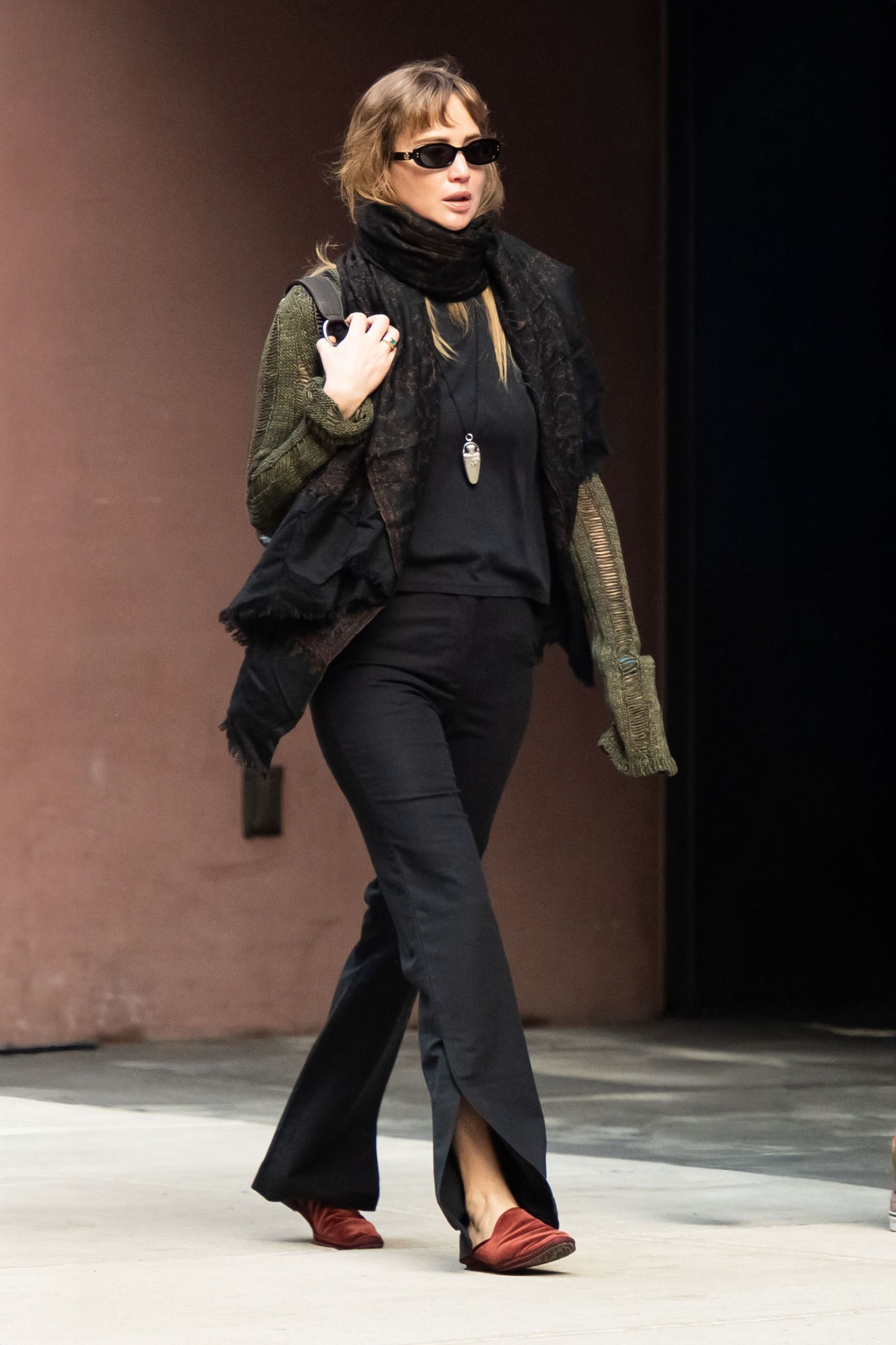 Jennifer Lawrence walks down the street in New York wearing black split-hem trousers with rust red satin shoes a black top, an oversized scarf and a green cardigan.