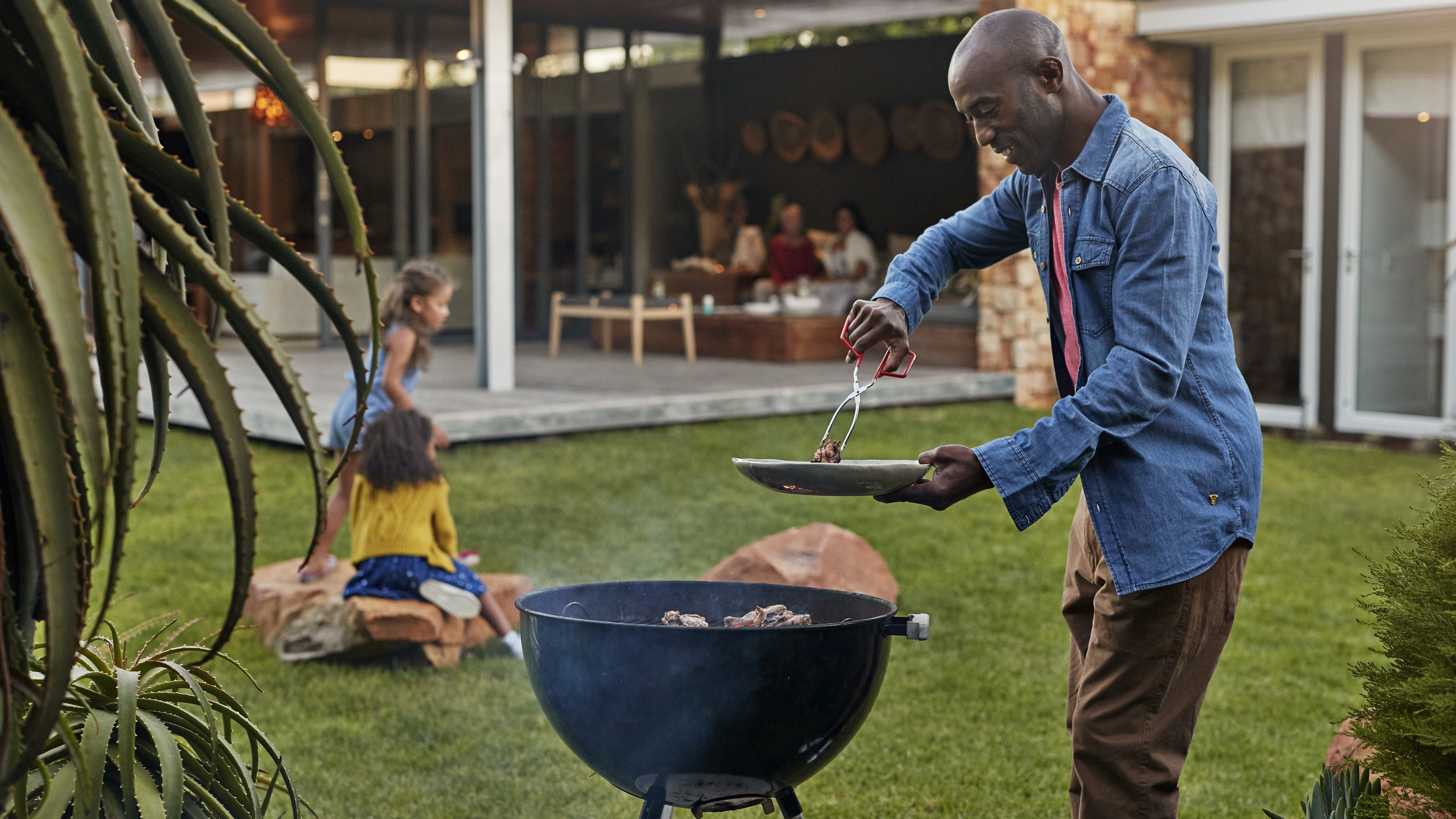 A man grills in his backyard as his daughters play nearby.