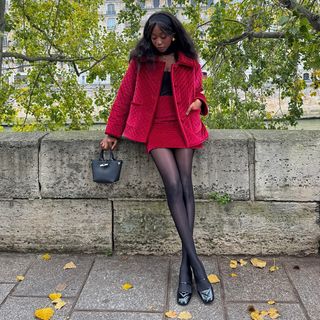 Emmanuelle is wearing a red velvet jacket, red velvet skirt, black top, black sheer tights, black ballet flats, black top-handle bag.