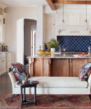 A kitchen with a white storage bench with a red rug underneath, natural and white wooden kitchen cabinets, and a marble kitchen island with yellow vases and strawberries on top of it