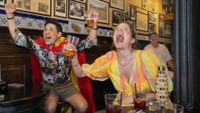 In a traditional Madrid bar, a man in a Spanish flag cape and a woman in a yellow and pink dress, both with red and yellow face paint, cheer ecstatically with beers raised as an older man smiles