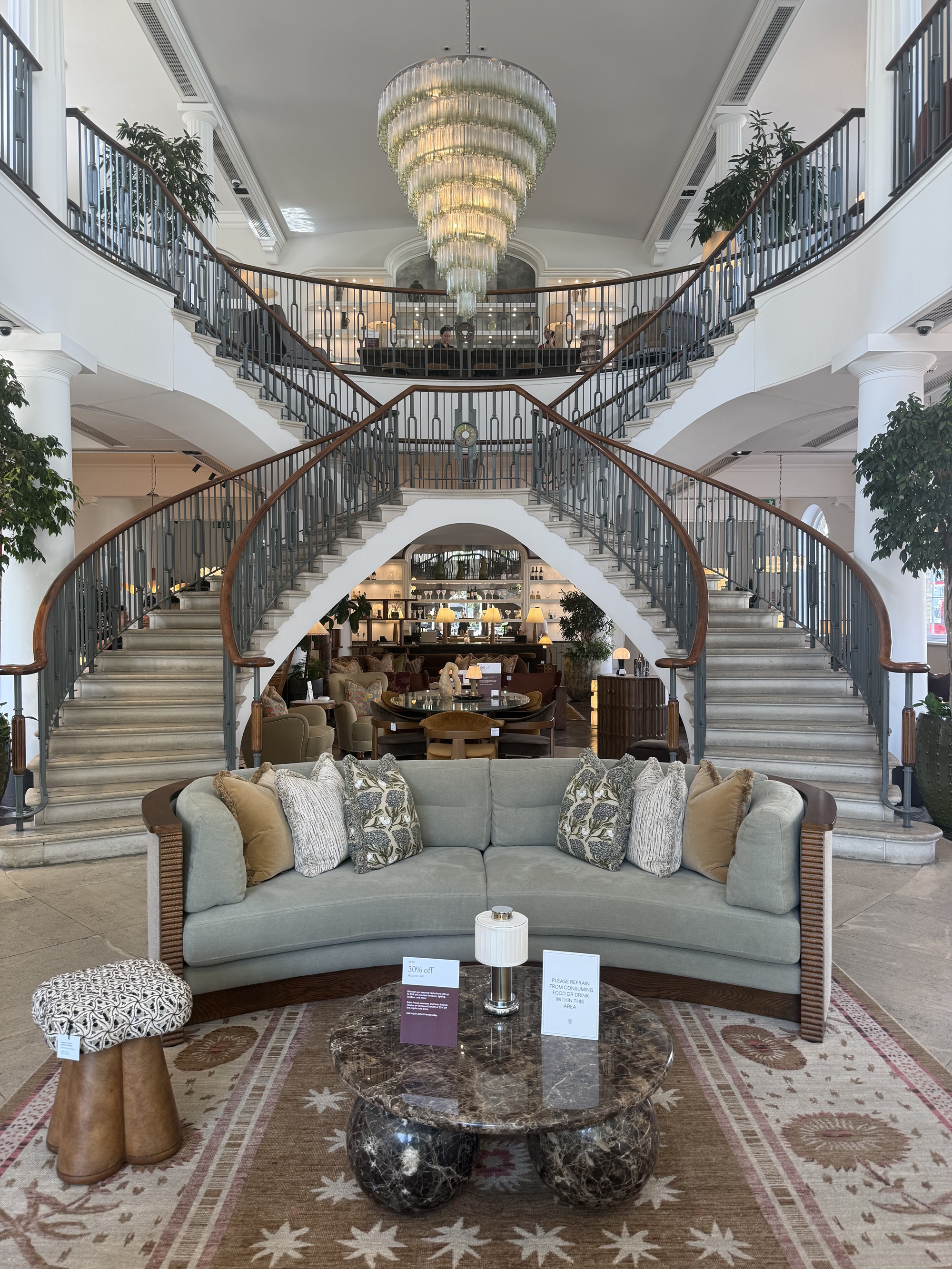soho home sloane showroom with grand staircase and chandelier, sofa with patterned rug, marble coffee table, ottoman, and cushions