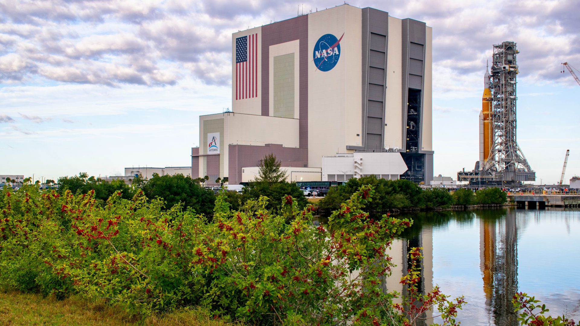 An orange rocket sits on a mobile scaffold as it rolls out of a large white building with the American flag and NASA meatball logo on the side.
