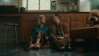 A press photo from Lessons in Chemistry of Brie Larson and Lewis Pullman sitting on the ground.