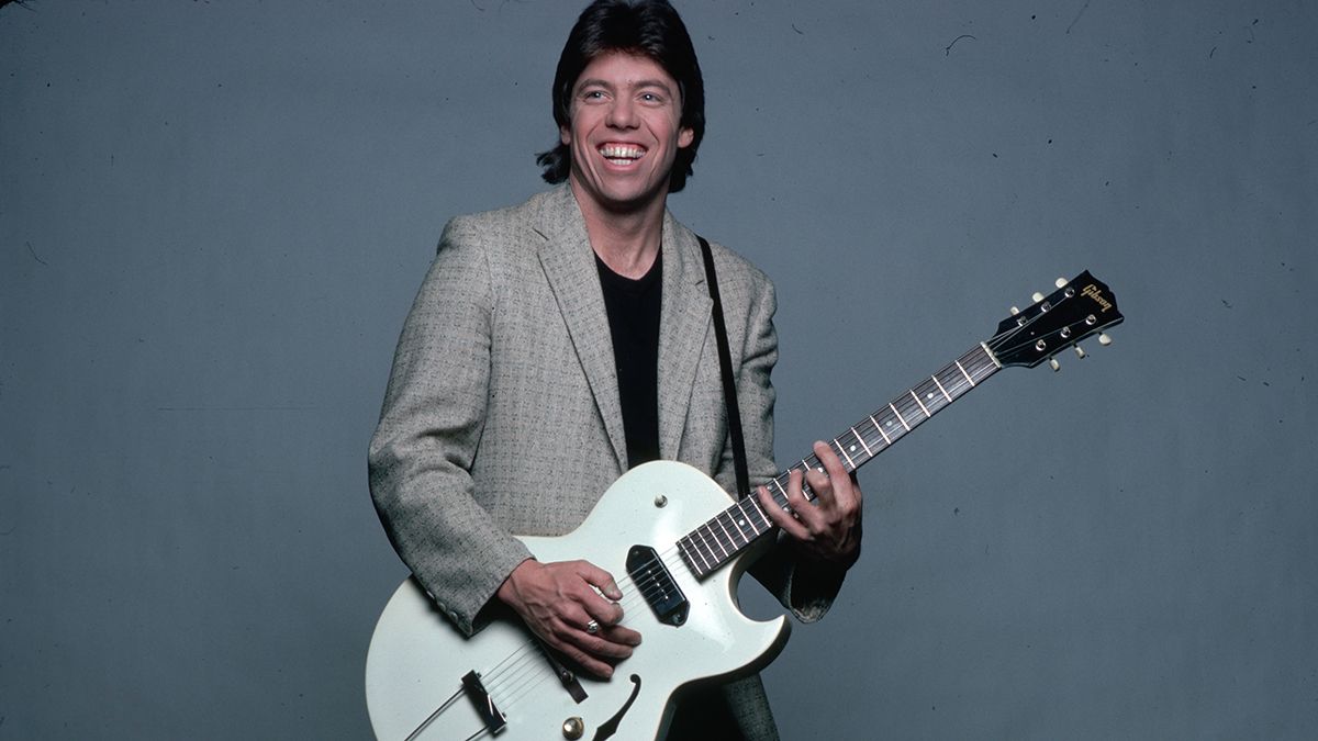 George Thorogood is shown in a studio portrait, wearing a tan jacket over a baclk t-shirt and pants. He is playing a guitar he has strapped on