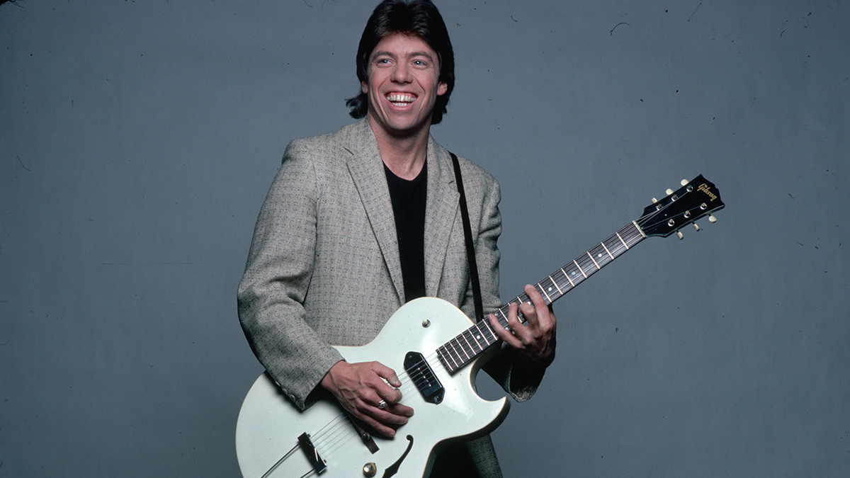 George Thorogood is shown in a studio portrait, wearing a tan jacket over a baclk t-shirt and pants. He is playing a guitar he has strapped on